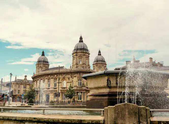 The Fountain in Queen’s Gardens, Hull, featuring a grand neoclassical building with three domes, elegant stone architecture, and decorative windows. A softly spraying fountain in the foreground reflects in calm water under a blue sky with clouds, creating a serene civic and historical scene.