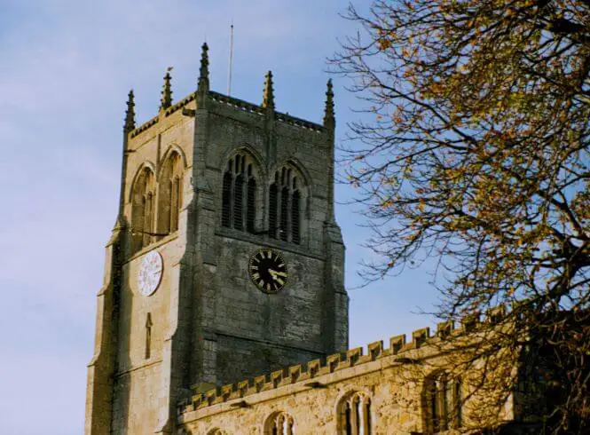 Historic stone church tower in Preston, Hull, illuminated by warm afternoon light, featuring Gothic-style arched windows, pointed battlements, and dual clock faces—one white and one black. A leafless tree with bare branches frames the tower against a soft sky, highlighting the contrast between timeless architecture and nature’s seasonal change in this tranquil East Yorkshire village scene.