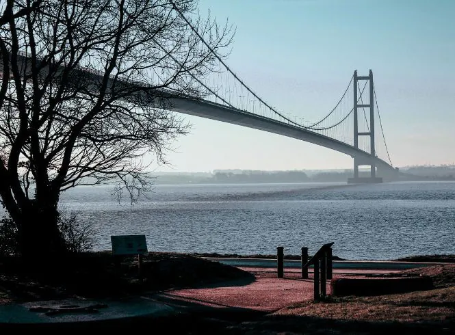 Scenic view of the Humber Bridge at Humber Bridge Country Park in Hessle, East Yorkshire, UK, featuring the elegant suspension bridge spanning calm blue water under a soft, cloudy sky. Leafless trees frame the foreground, their dark branches contrasting with the tranquil river and light horizon. The image captures the bridge’s towering structure and sweeping cables, symbolizing engineering achievement and connection amid a peaceful natural landscape during late autumn or winter.