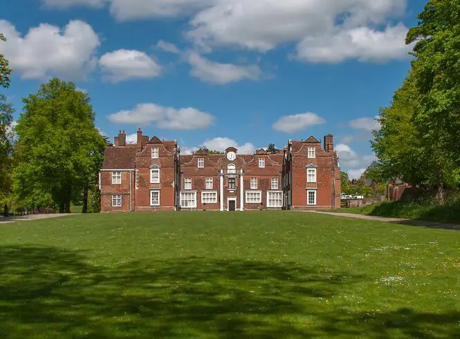 Christchurch Mansion, Ipswich, a grand red-brick mansion with white window frames and a central clock, set amid a sprawling green lawn and framed by lush trees under a bright blue sky with scattered white clouds, evoking serene and timeless elegance