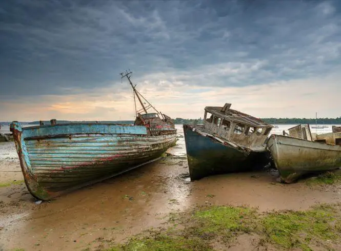 Two abandoned, weather-beaten fishing boats resting on the muddy shore of the River Orwell at Pin Mill during low tide, with peeling paint and broken cabins, surrounded by patches of vegetation under a soft blue and orange sky at dawn or dusk.