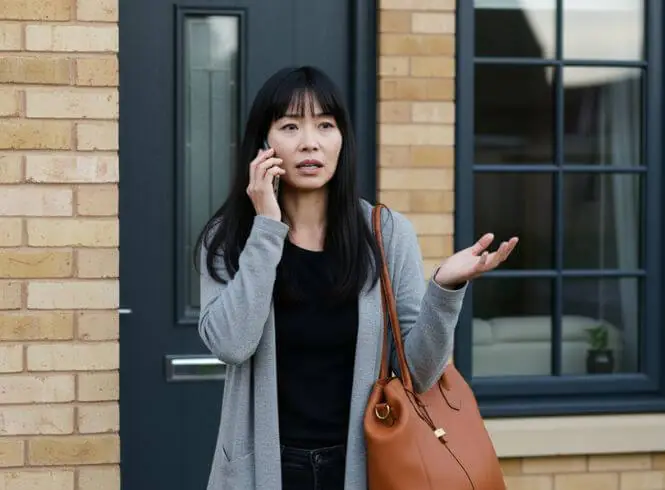 Woman locked out of her home calling a locksmith, standing by a brick exterior and dark front door. She looks concerned while holding her phone, with a raised hand gesture and a brown handbag over her shoulder.
