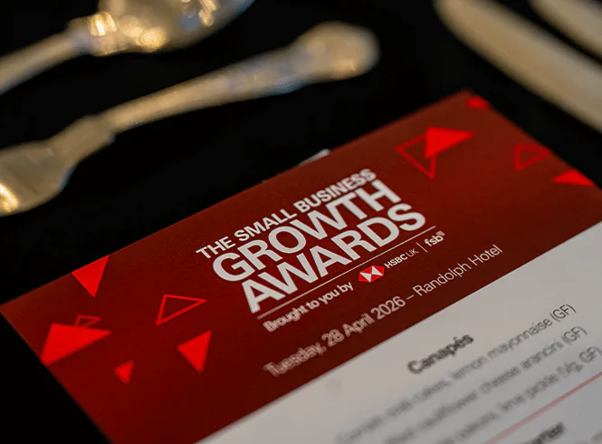 Close-up of a red and white Small Business Growth Awards invitation dated Tuesday, 28 April 2026, with silverware in the background.